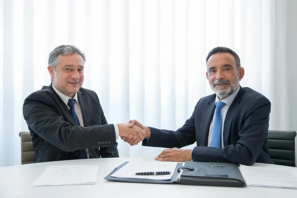 Two business partners in suits shaking hands over company documents in a Thai office meeting